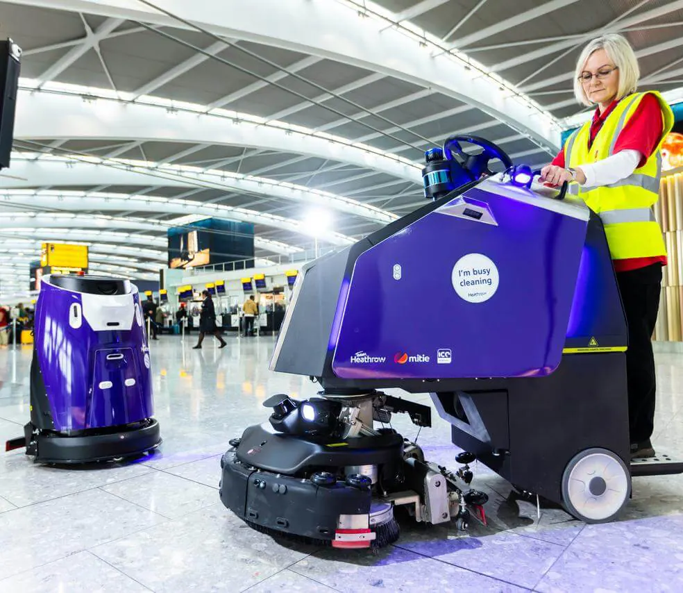 Gausium Scrubber 75 : Robot Gausium habillé en violet avec une opératrice à l'aéroport de Heathrow (984×854) ; modèle probablement Scrubber 75 mais non certain, habillage client.